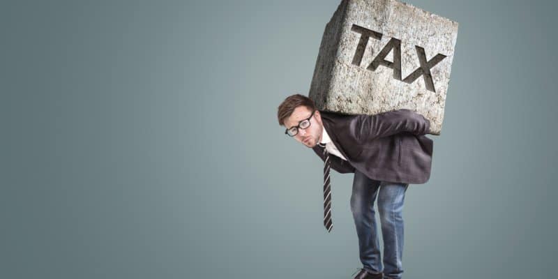 Concept of a businessman bending under the burden of high taxes. He is carrying a large stone on his back with the word "TAX" printed on it.